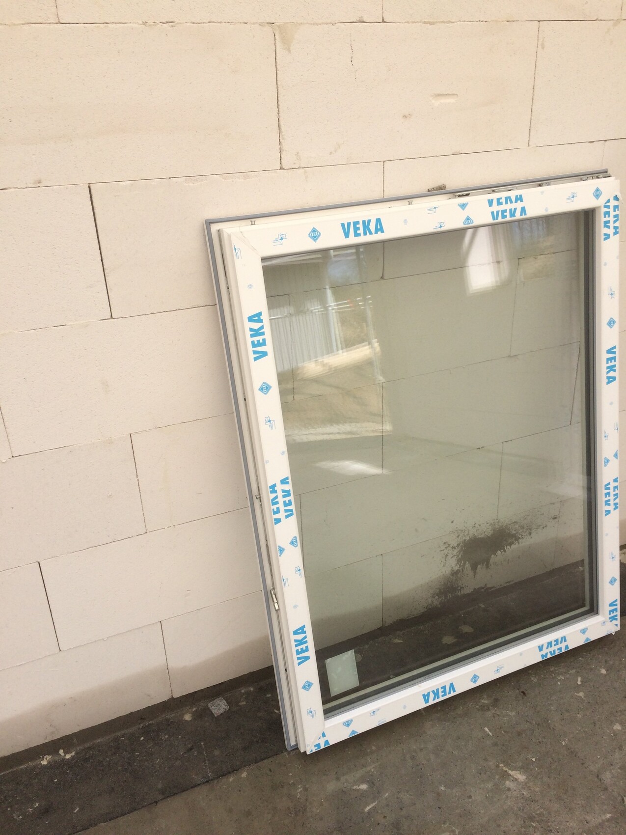 Großes Fenster mit Glas lehnt an einer Steinwand; Rahmen mit VEKA-Schutzfolie.
