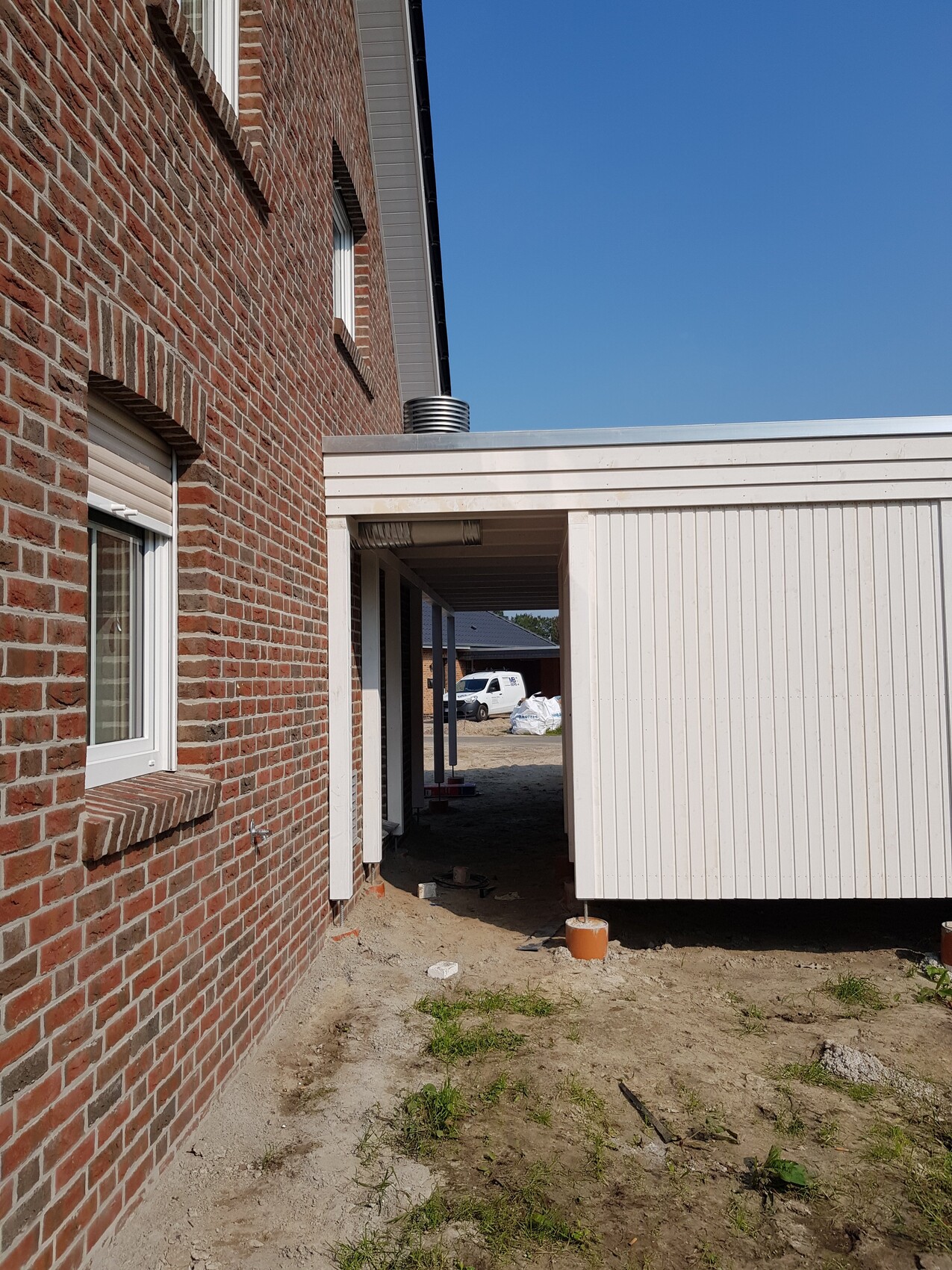 Außenansicht eines roten Backsteinhauses mit weißem Anbau und Baumaterial auf Sandboden.