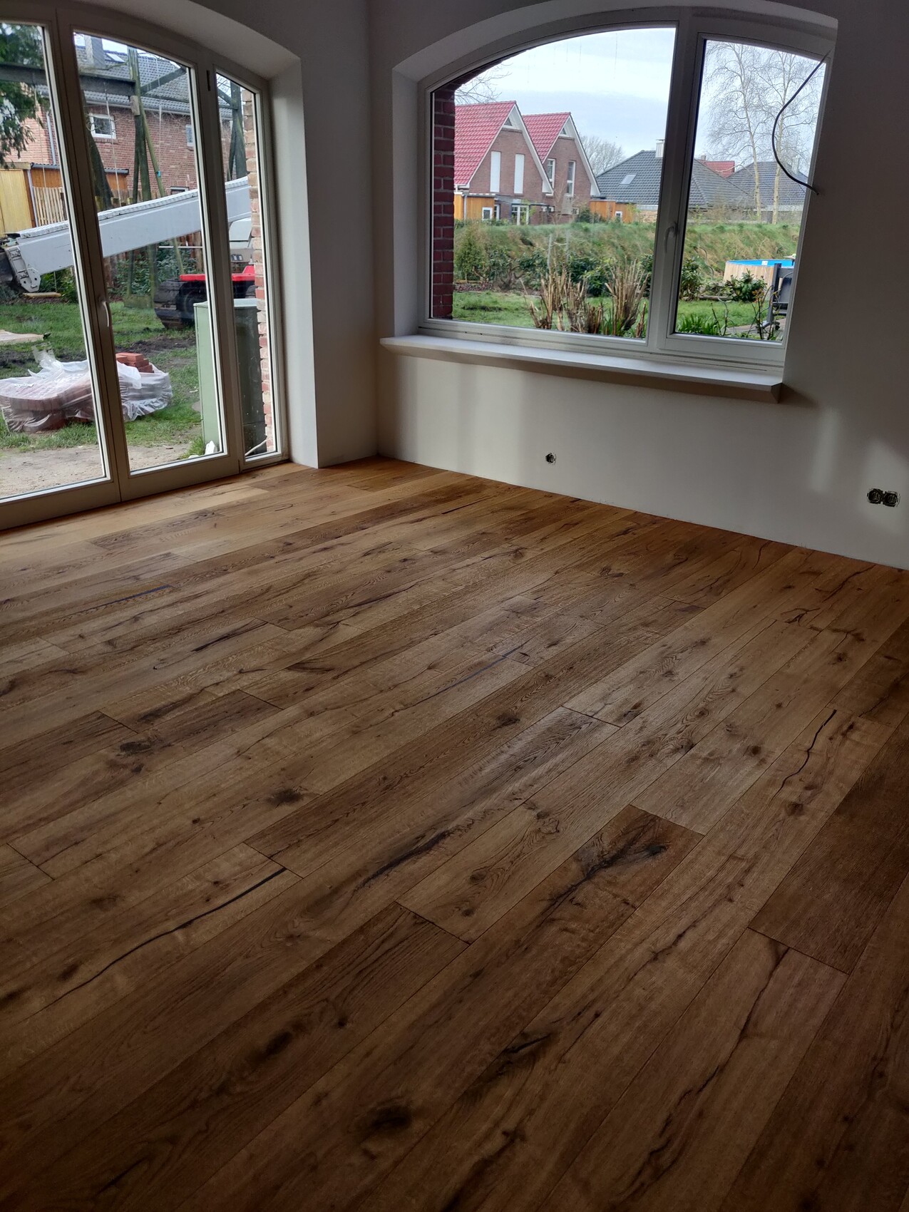 Leerer Raum mit Holzboden, großer Fensterfront und Blick nach draußen auf Garten.