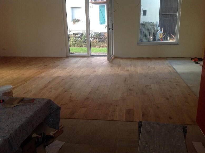 Wohnzimmer mit halbfertigem Holzboden, Glas-Schiebetür zum Garten, Werkzeuge links.