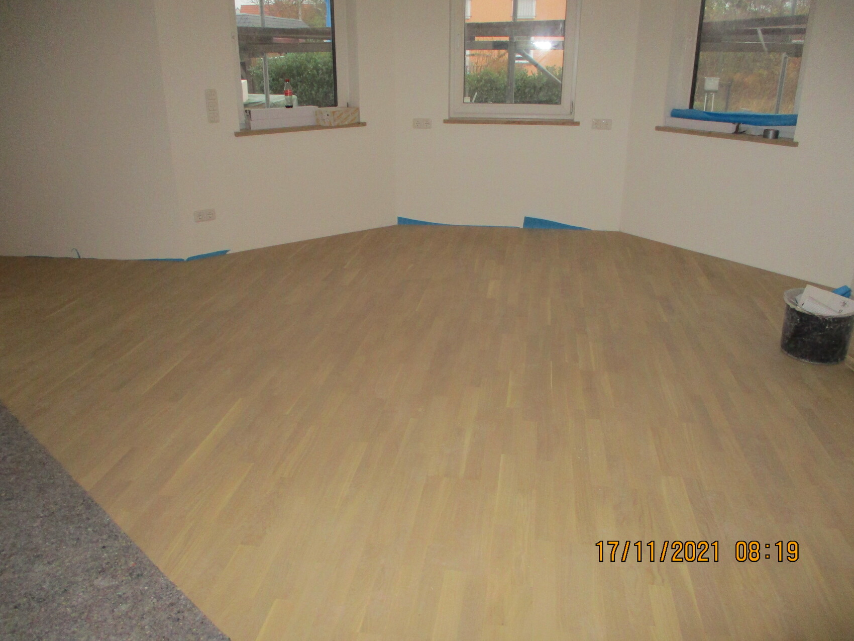 Leerer Raum mit neu verlegtem Holzboden, blaue Abdeckbandkanten an Wänden, Eimer rechts