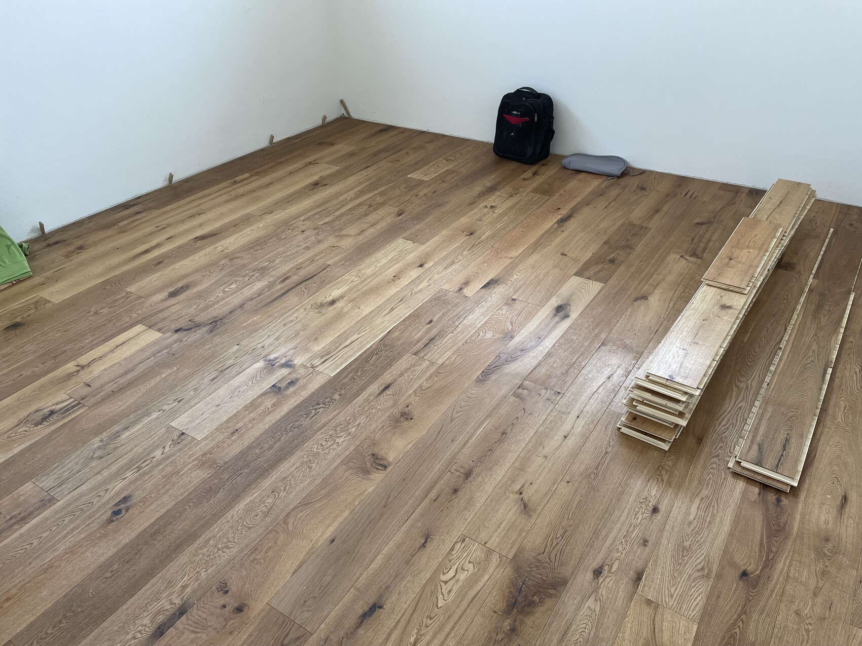 Neu verlegter Holzboden, rechts ein Stapel Holzdielen, schwarzer Rucksack in der Ecke.