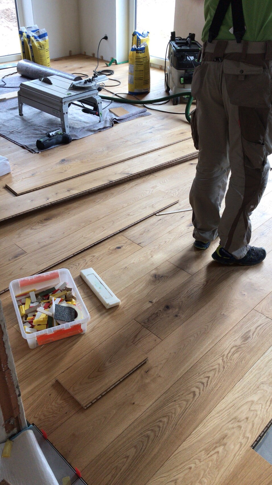 Renovierungsraum mit Holzdielen, Kappsäge, Kabeln, gelben Säcken; Arbeiter steht rechts.
