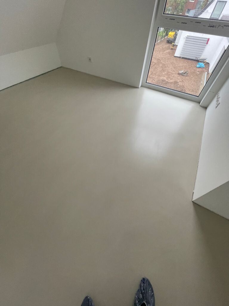 Leerer Raum mit hellem Boden, weiße Wände und großes Fenster zur Außenbaustelle.