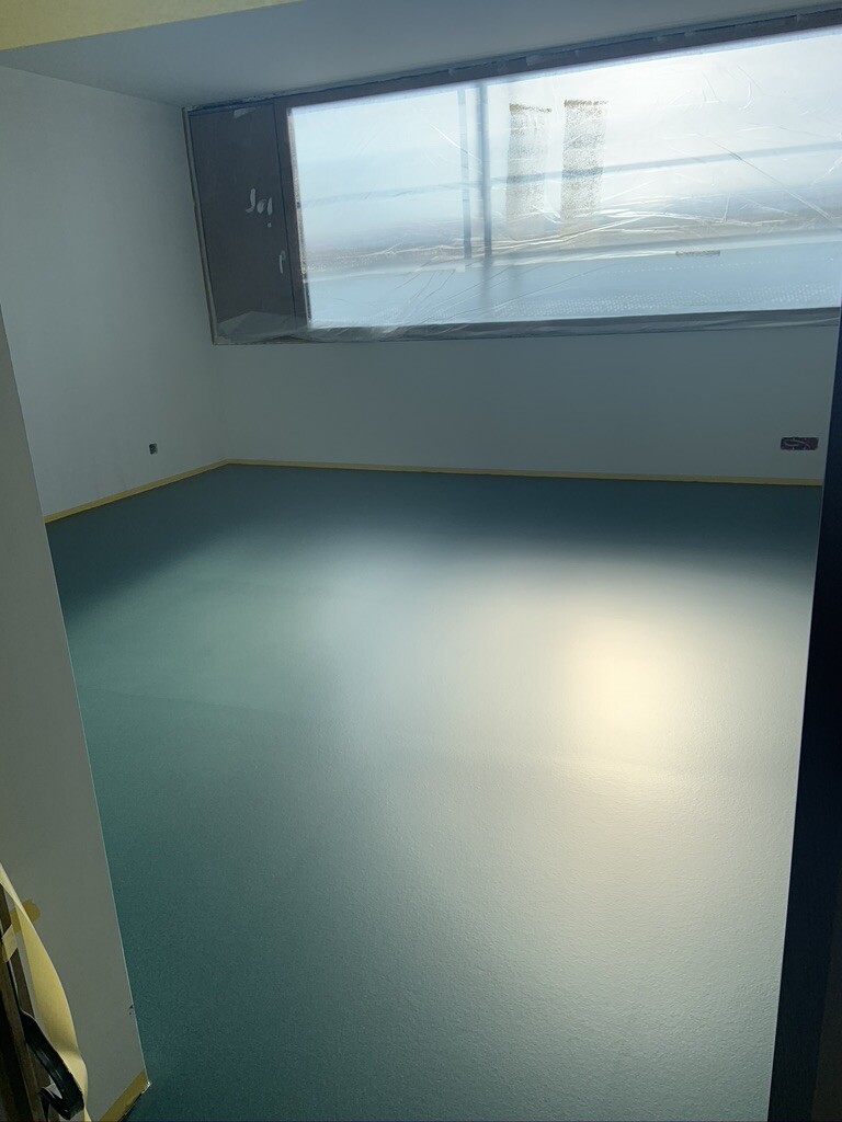 Leeres Zimmer mit türkisfarbenem Boden, weißen Wänden, großem Fenster mit Schutzfolie.