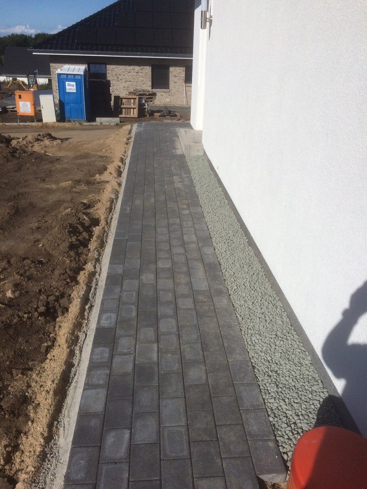 Gepflasterter Gehweg aus grauen Steinen neben weißer Hauswand; Baugeräte im Hintergrund.