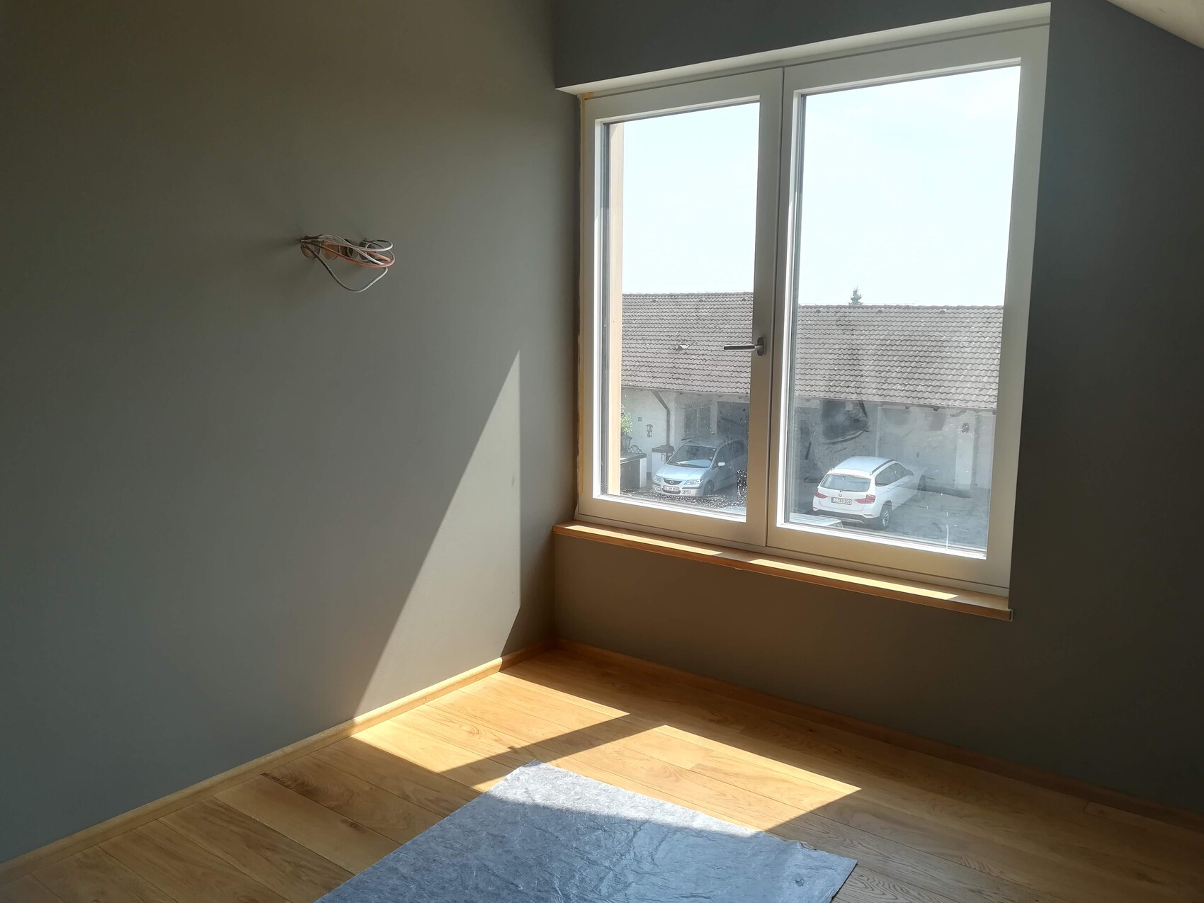 Leerer Raum mit grauen Wänden, Kabelbündel an der Wand, Fenster mit Blick auf Autos, Holzboden.