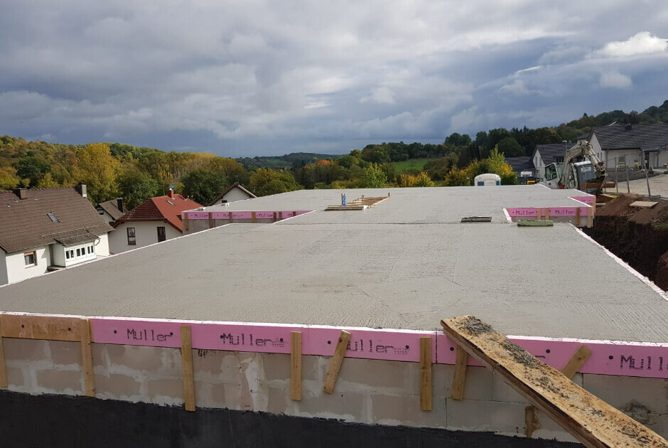Baustelle: Flachdach mit pinken Dämmplatten am Rand, Herbstwald und Häuser im Hintergrund.