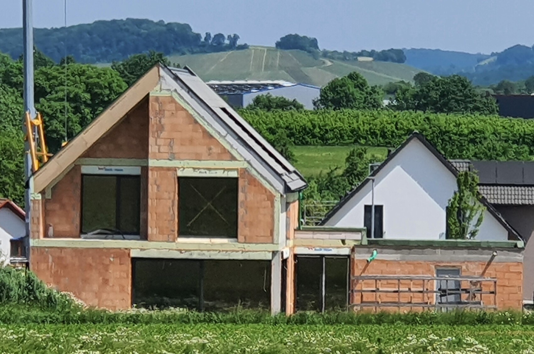 Unfertig gebautes Backsteinhaus mit großen Fenstern, daneben ein weißes Haus; grüne Felder dahinter.