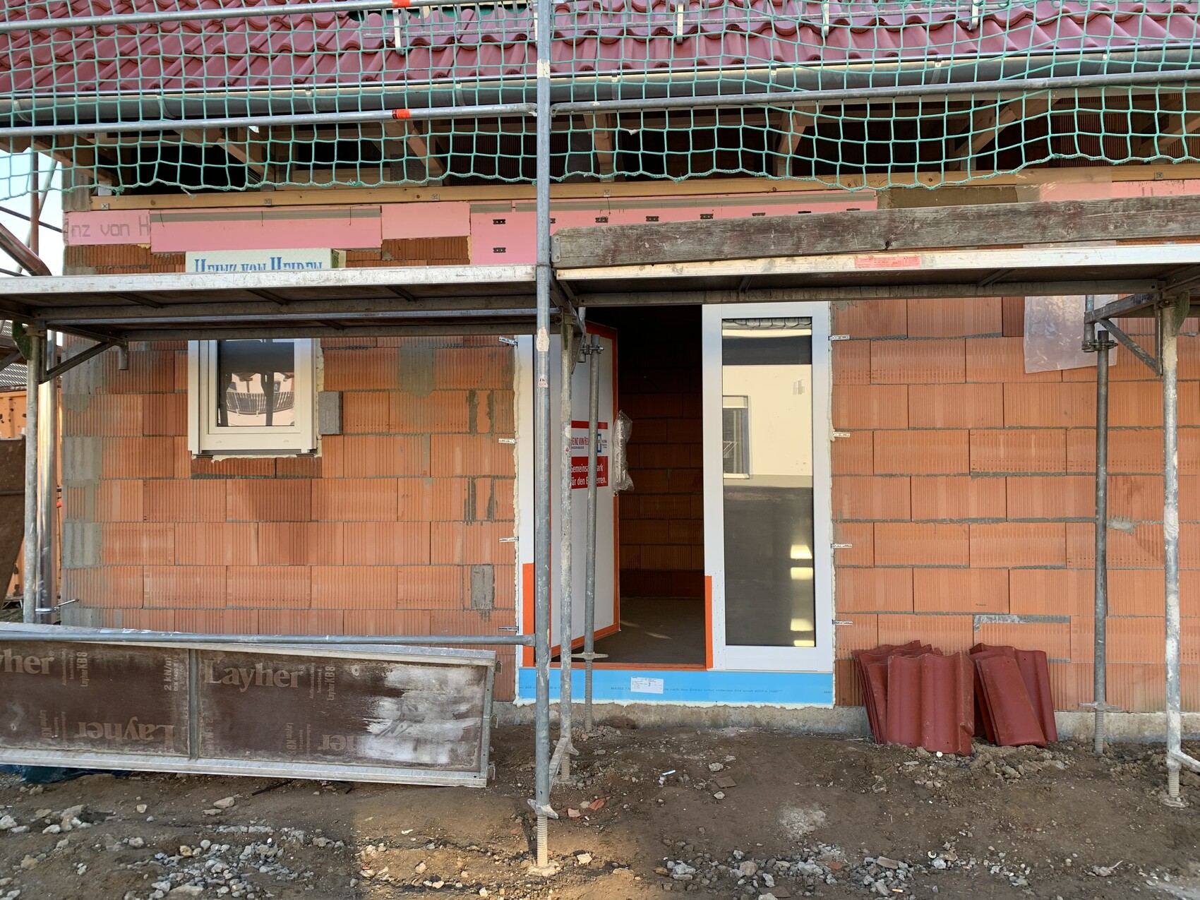 Baustelle vor rotem Ziegelbau, offene Tür mit Glas, linkes Fenster, Gerüst und Schutt.