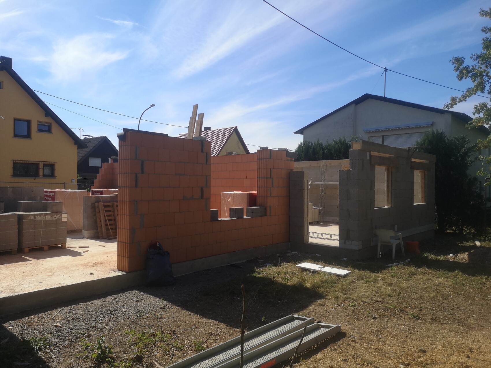 Rohbau aus roten Ziegeln mit unfertigen Wänden auf einer Baustelle, Häuser im Hintergrund.