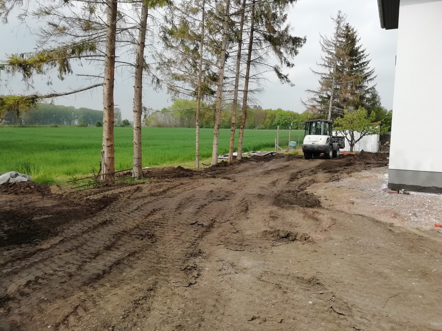 Unbefestigter Erdweg neben grünen Feldern, links Baumreihe, rechts Baufahrzeug vor weißem Gebäude.