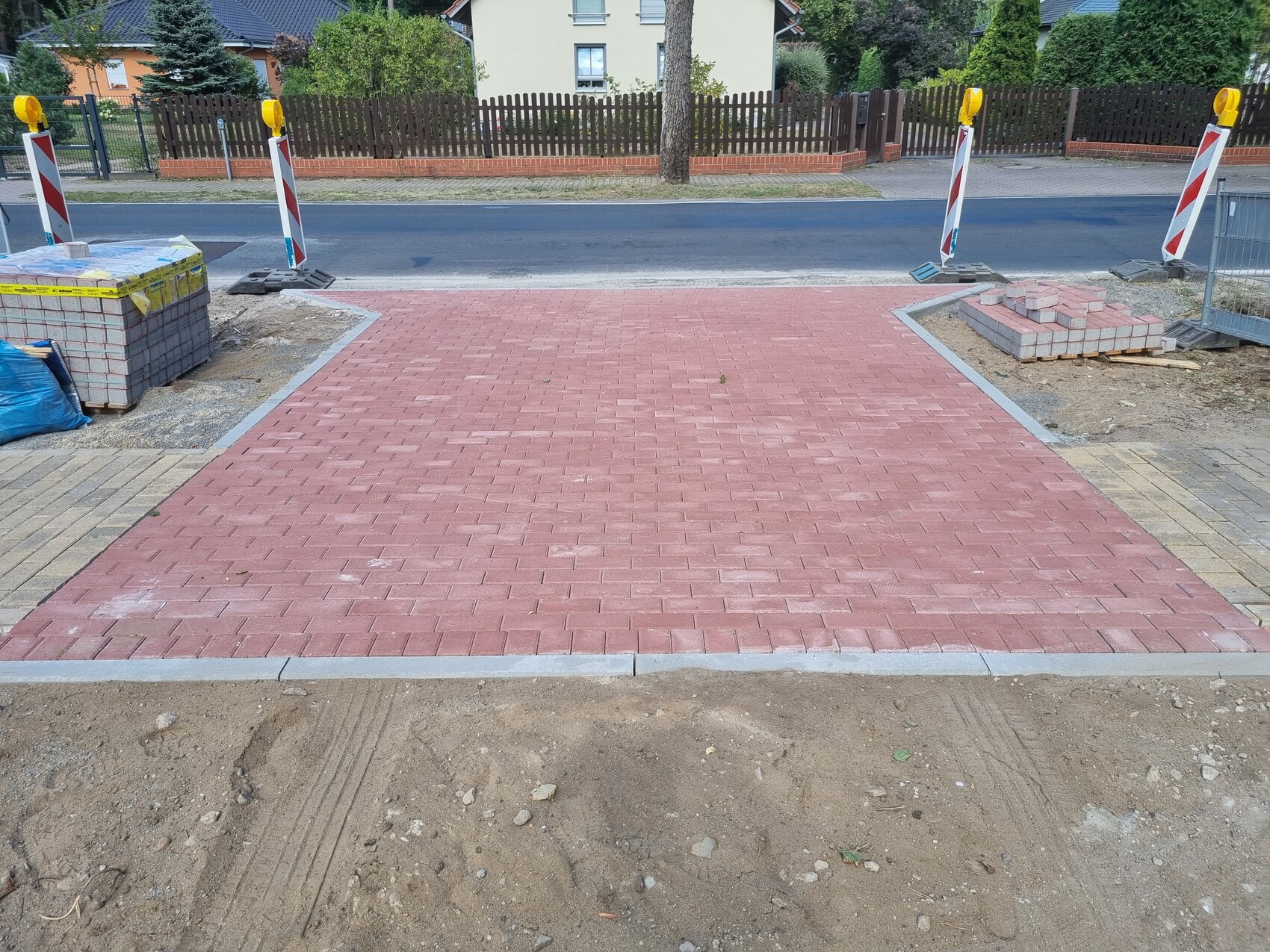 Rot gepflasterter Gehweg aus Betonsteinen führt zur Straße; Baustellenabsperrungen links und rechts.