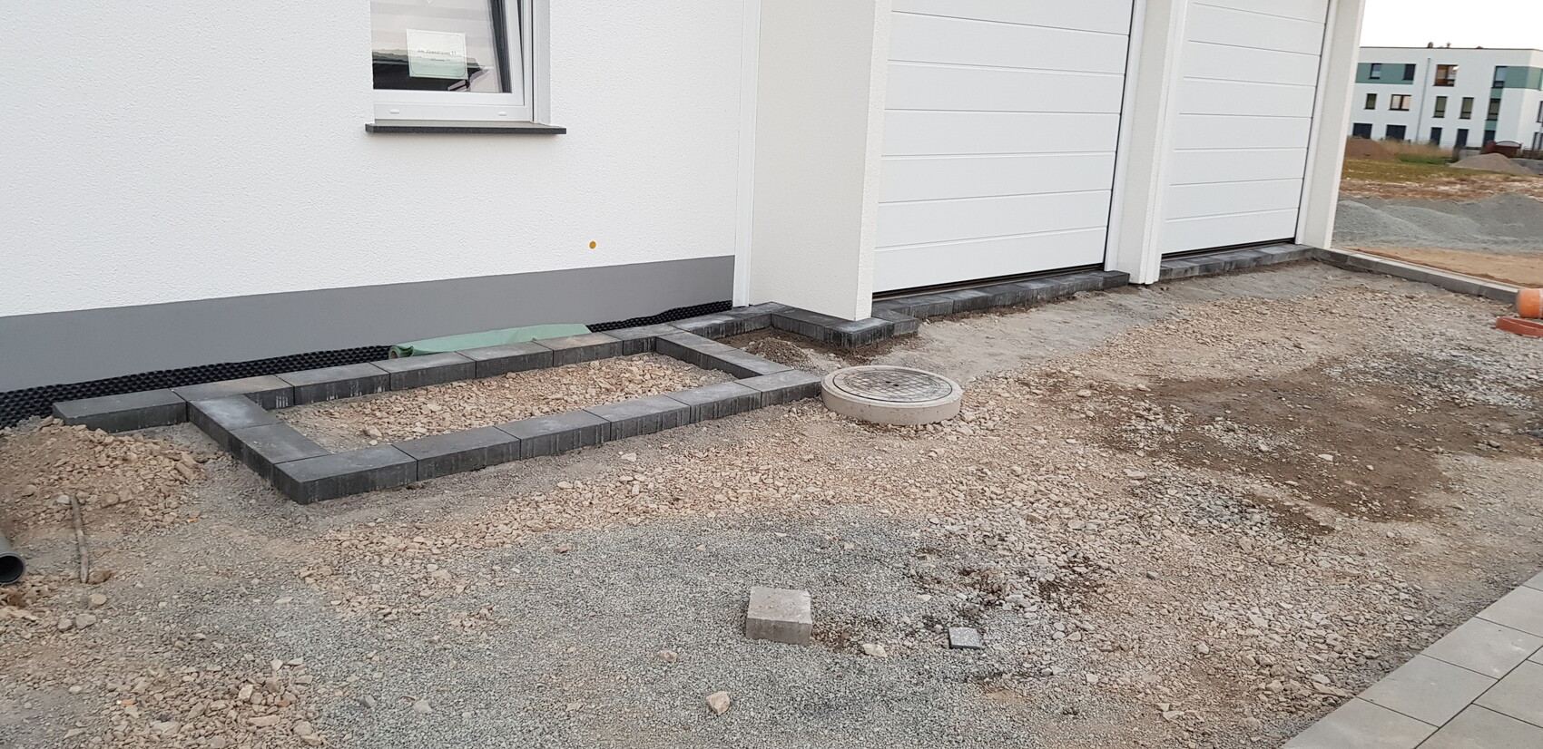Außenansicht: Pflasterrahmen am Haus, Kiesboden, runde Abdeckung, Garage im Hintergrund.