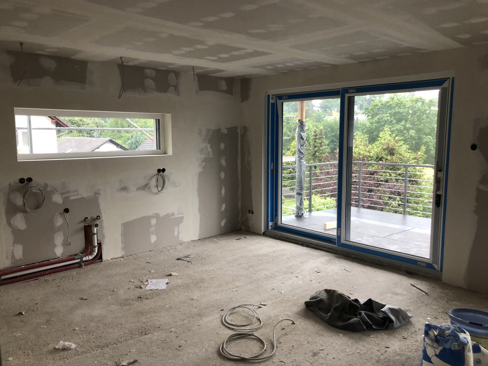 Unfertiger Renovierungsraum mit freiliegenden Kabeln und Rohren und Balkon-Schiebetür zum Außenbereich.