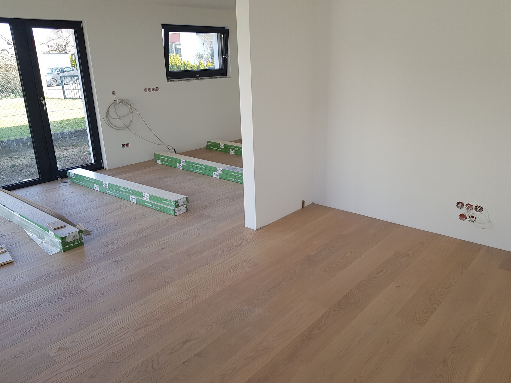 Renovierungsraum mit hellem Laminat, verpacktem Bodenmaterial, Kabeln an der Wand, linke Tür.