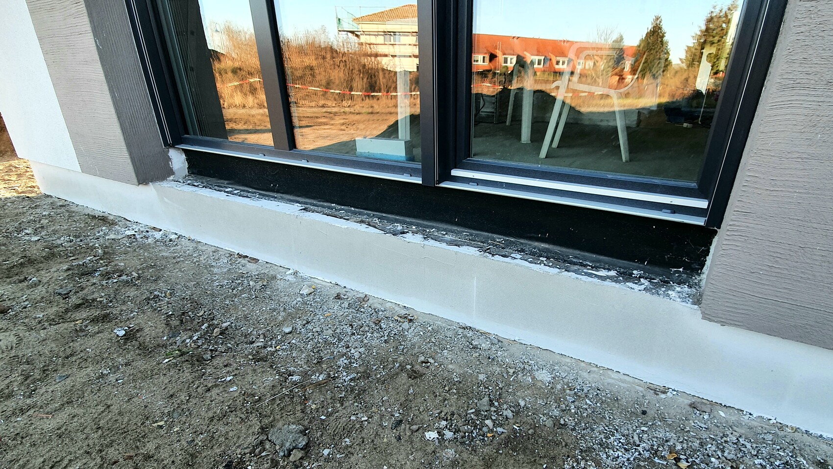 Außenansicht eines Neubauhauses: dunkle Fensterbank, weißer Sockel, unbefestigtes Erdreich davor.
