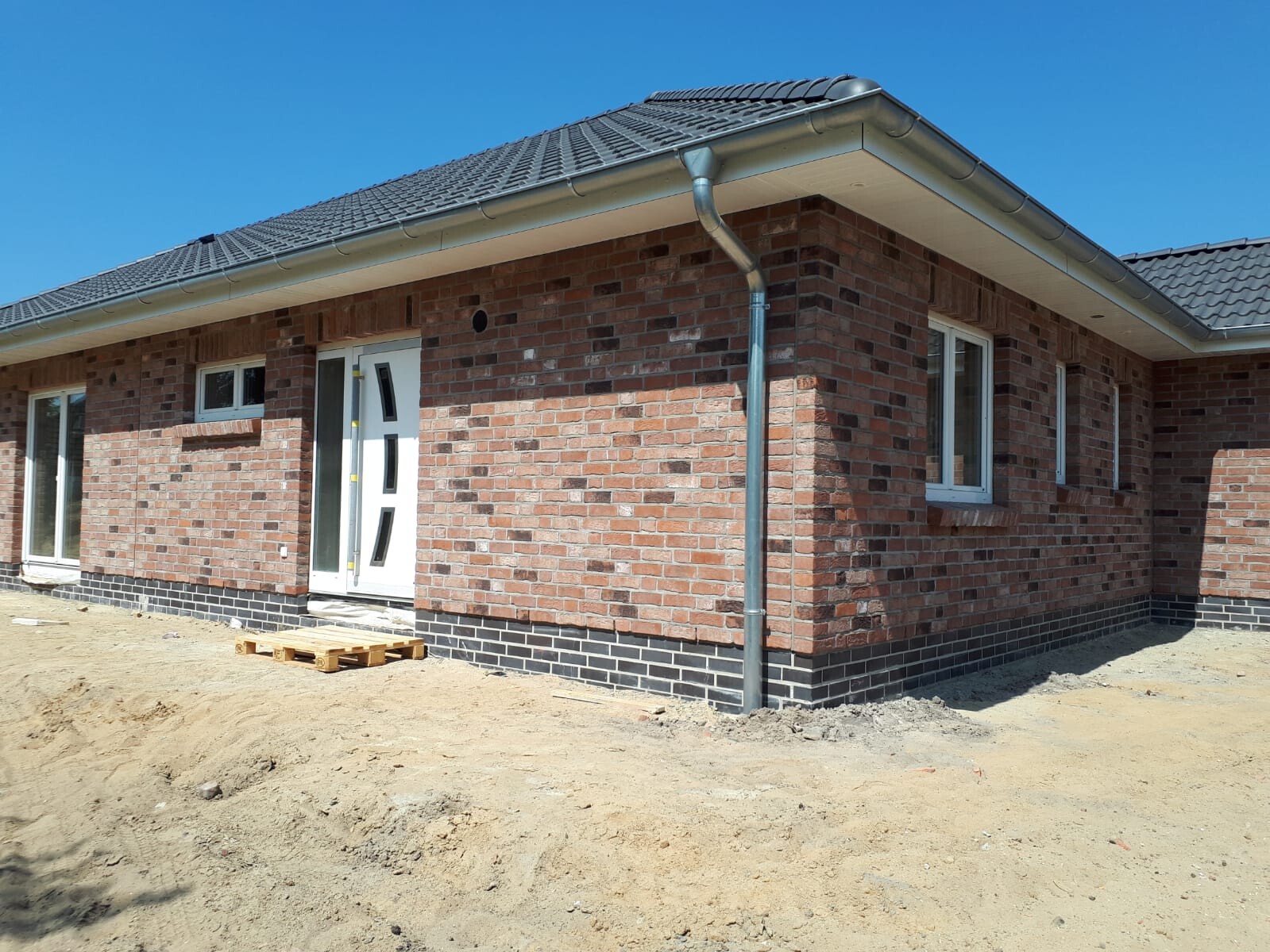 Neubau aus rotem Backstein mit weißen Fenstern, Dachrinne und Baugrund davor.