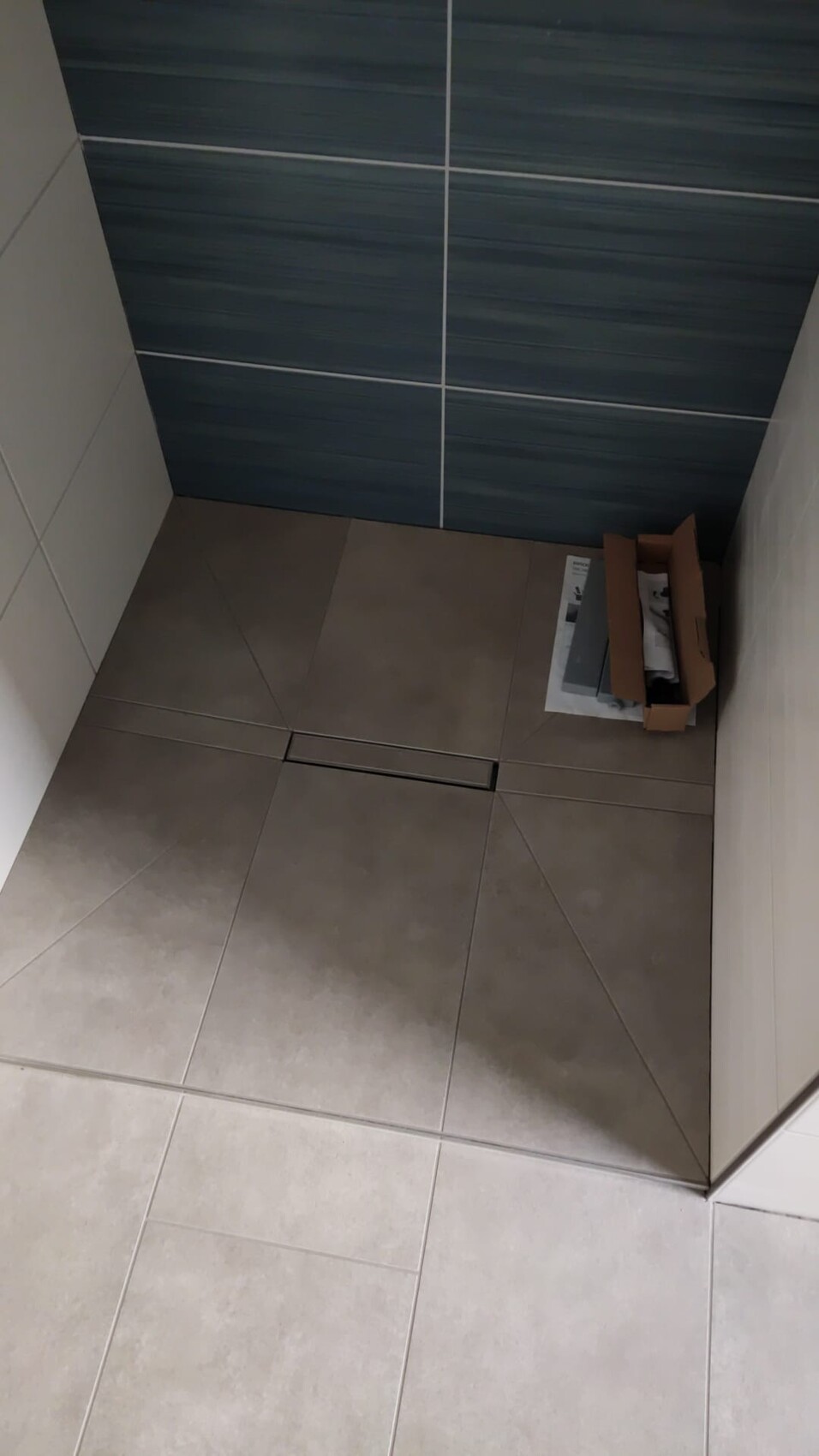 Eckbad: beige Bodenfliesen, blau gestreifte Wandfliesen, lange Bodenrinne, Karton daneben