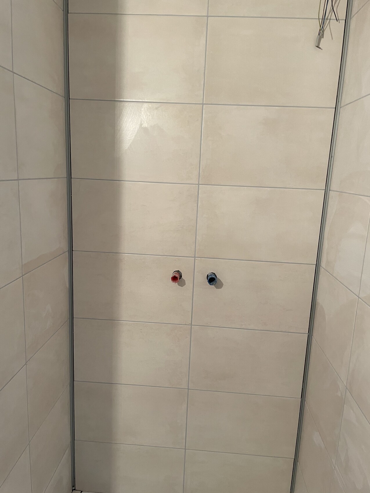 Duschkabine mit hellen Wandfliesen, zwei Wasseranschlüsse (rot/blau) an der Wand.