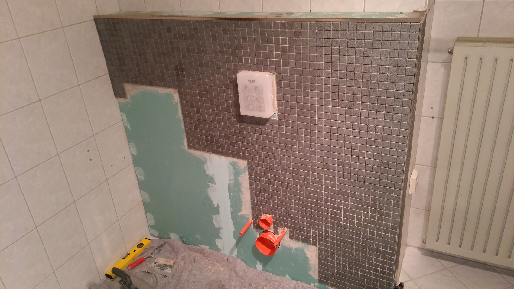 Badezimmerwand in Renovierung: dunkelgraue Mosaikfliesen, roter Rohranschluss, grüne Wandfläche.