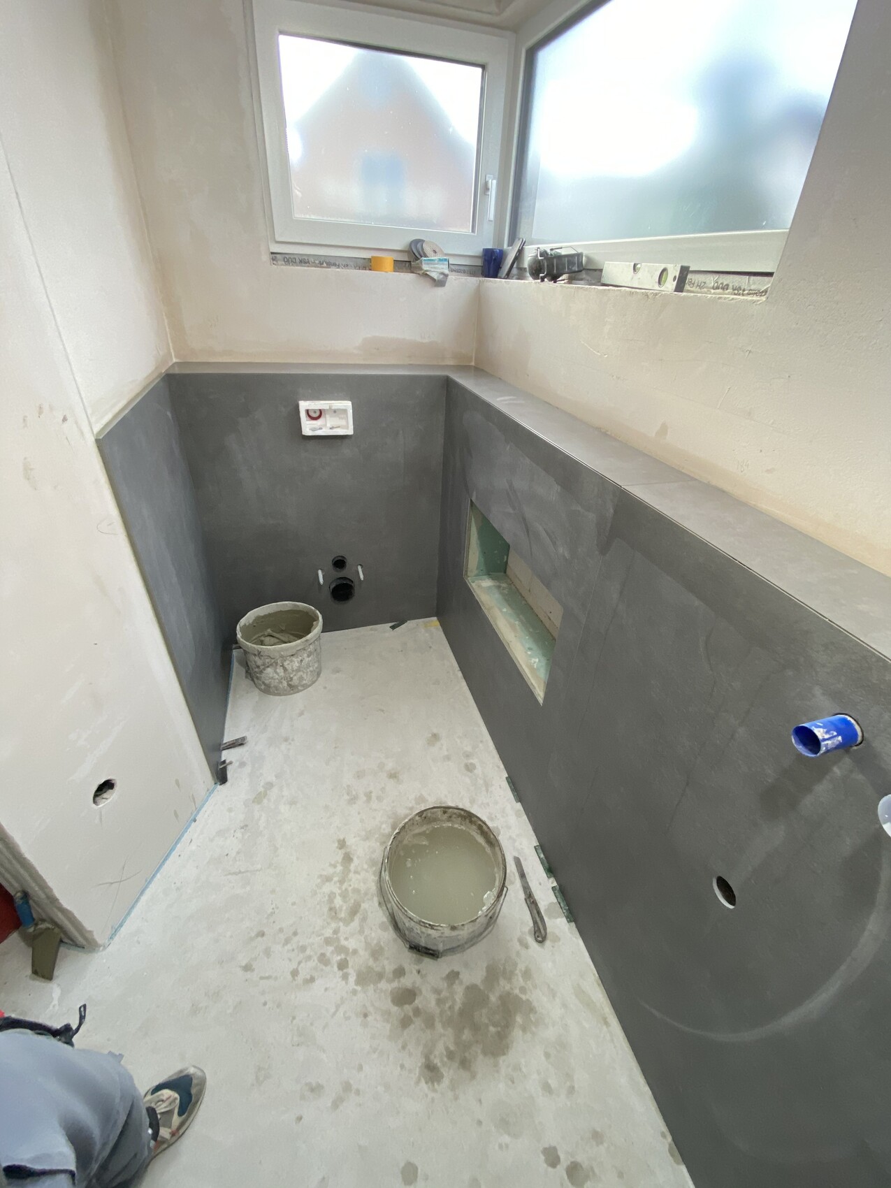 Kleines Badezimmer im Renovierungszustand; graue Wandverkleidung, Fenster, Eimer mit Mörtel, Rohre sichtbar.