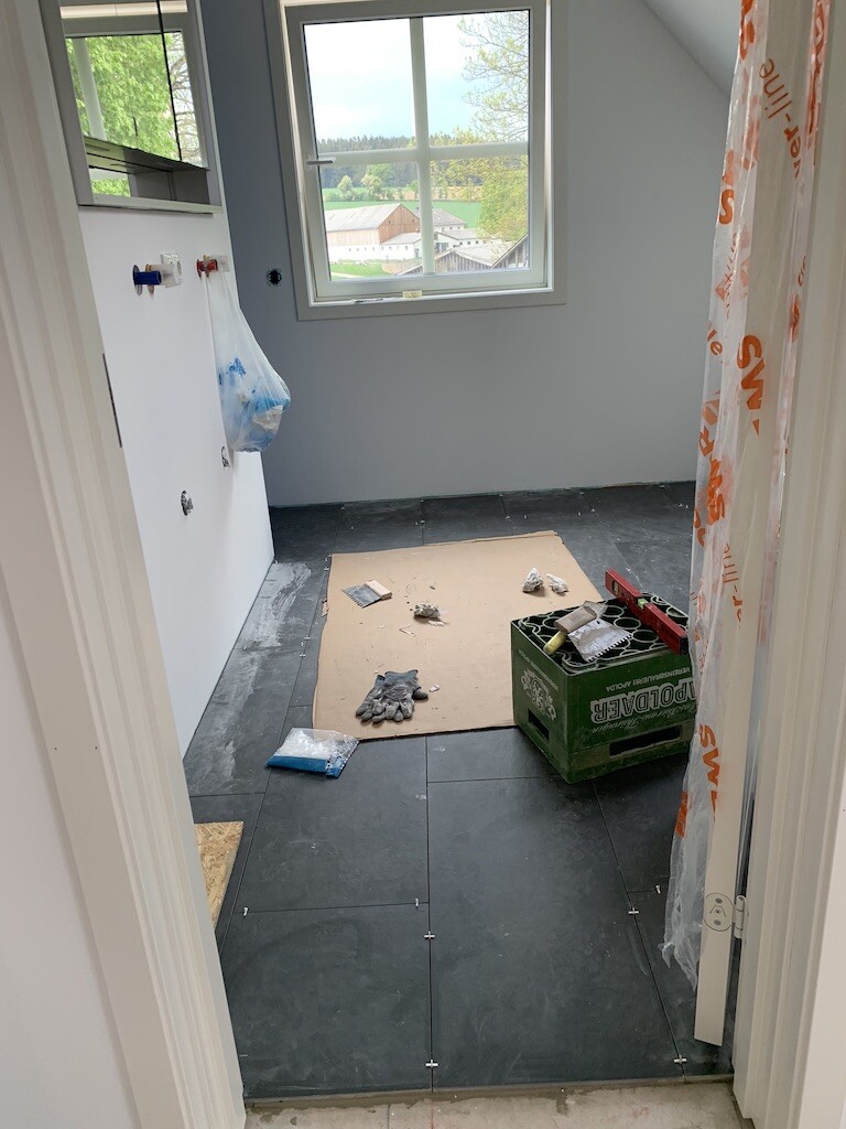 Badezimmer im Bau mit dunklen Fliesen, Werkzeugkiste, Handschuhe, Fenster mit Gartenblick.