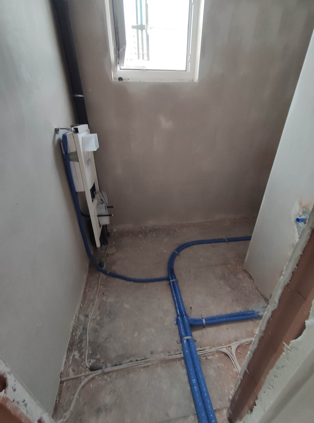 Kleiner, unfertiger Sanitärraum mit blauen Rohrleitungen auf dem Boden und Fenster in der Wand.