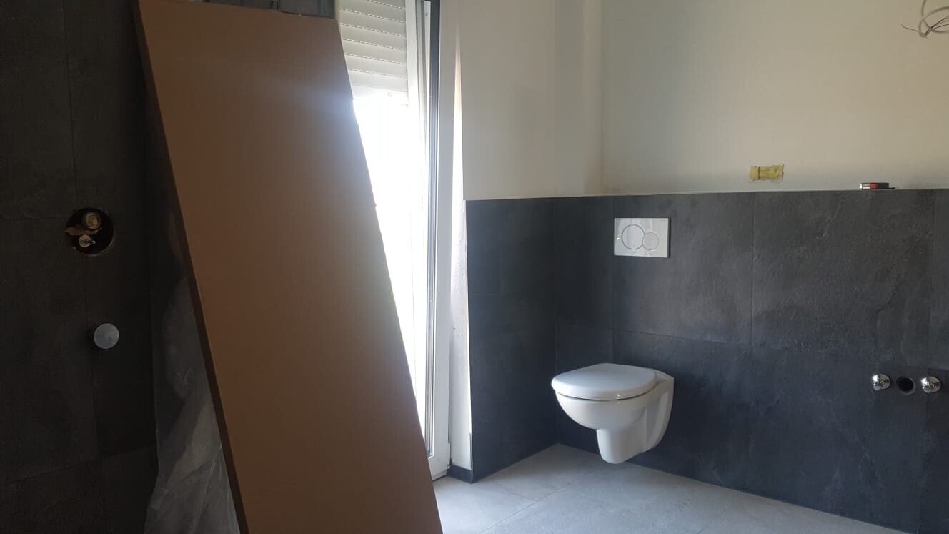 Modernes Badezimmer im Bau: Wand-WC, dunkle Fliesen, unfertige Installationen, Fenster links.