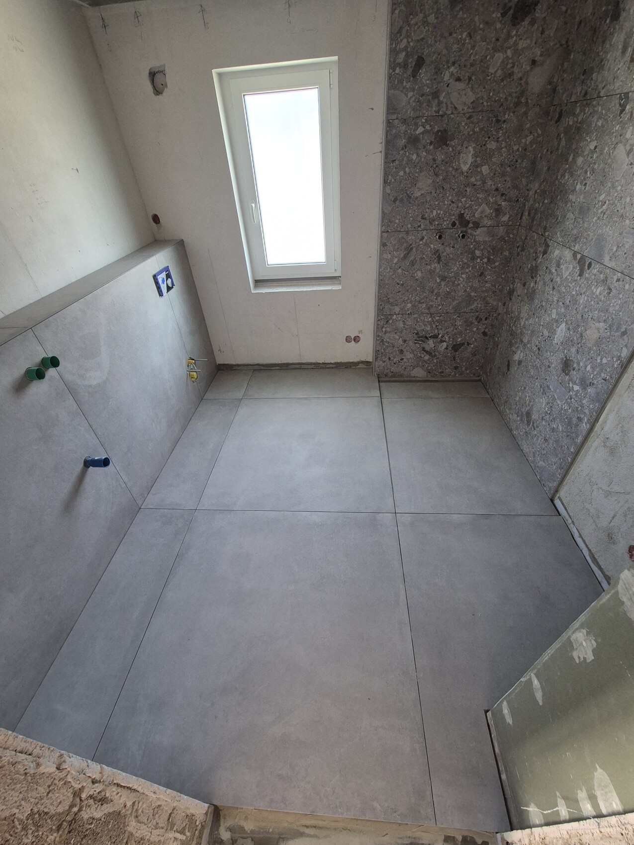Badezimmer im Rohbau mit großen grauen Bodenfliesen, Fenster und sichtbaren Sanitärleitungen.