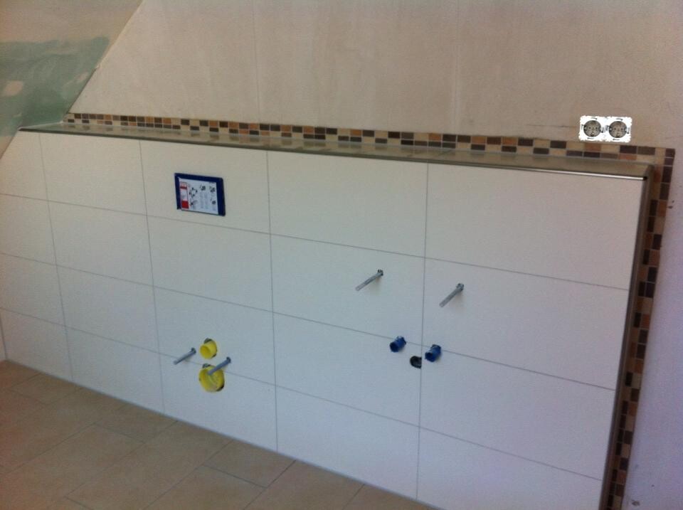 Weißer Bad-Unterschrank mit zwei Türen, sichtbaren Wasseranschlüssen (gelb/blau) und Fliesenboden.