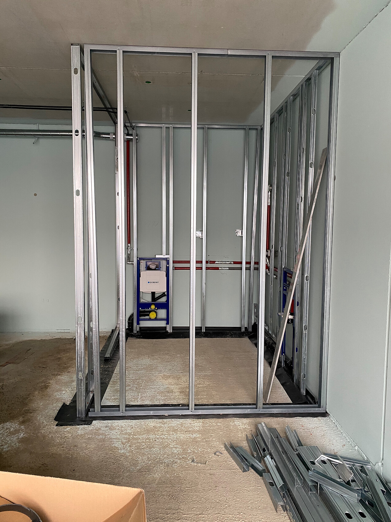 Metallständer-Wandrahmen auf Baustelle, rote Rohrleitungen, blaue Installationsstation im Hintergrund.
