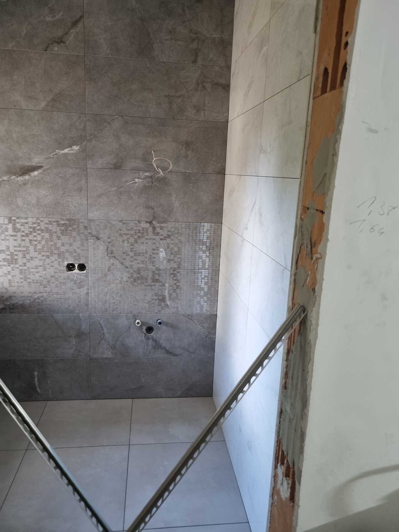Badezimmer im Bau: graue Wandfliesen, Mosaikband, Rohranschlüsse, offener Wandbereich rechts.