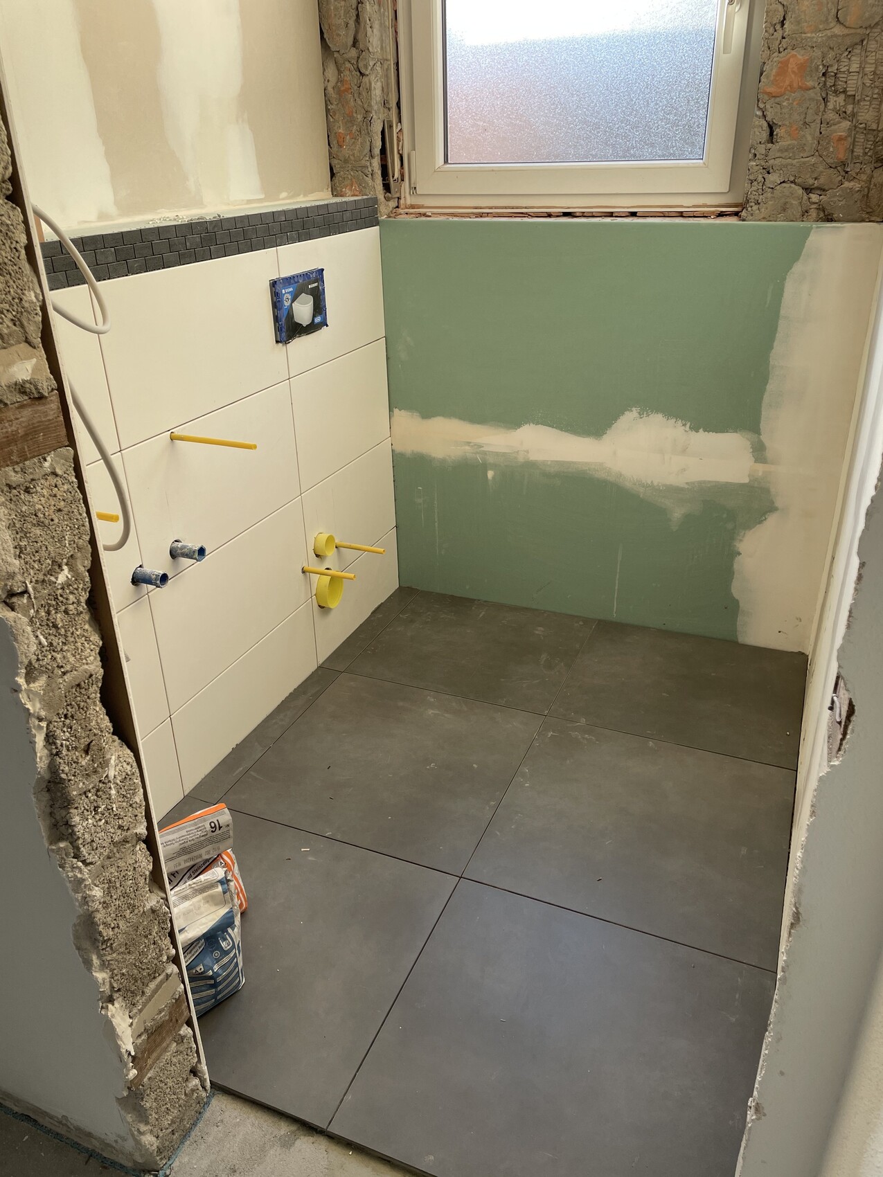 Badezimmer im Umbau: graue Bodenfliesen, weiße Fliesen, gelbe Rohre, grüne Wand, Fenster.
