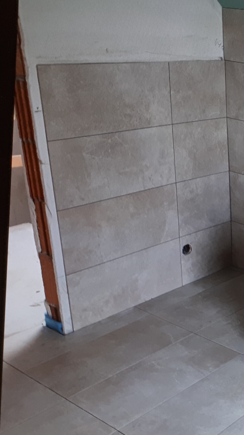 Innenraum mit grauen Wand- und Bodenfliesen; Wandloch für Sanitäranschluss, Baufortschritt sichtbar.