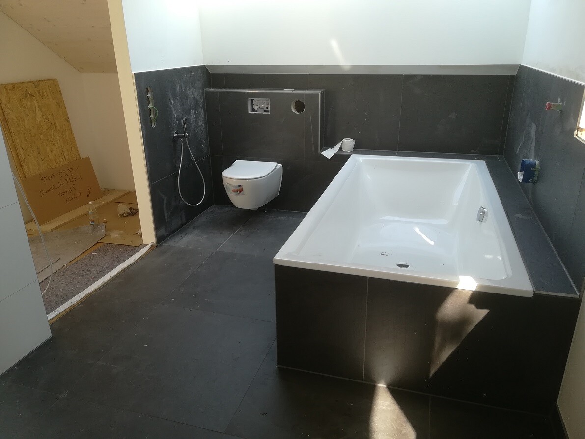 Modernes Bad im Bau: dunkle Fliesen, eingelassene Badewanne, wandhängendes WC, Bidet-Spray.