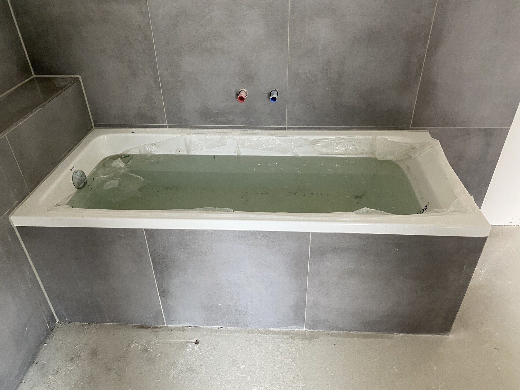 Moderne rechteckige Badewanne, graue Fliesen; Schutzfolie in der Wanne, rot/blau Anschluesse