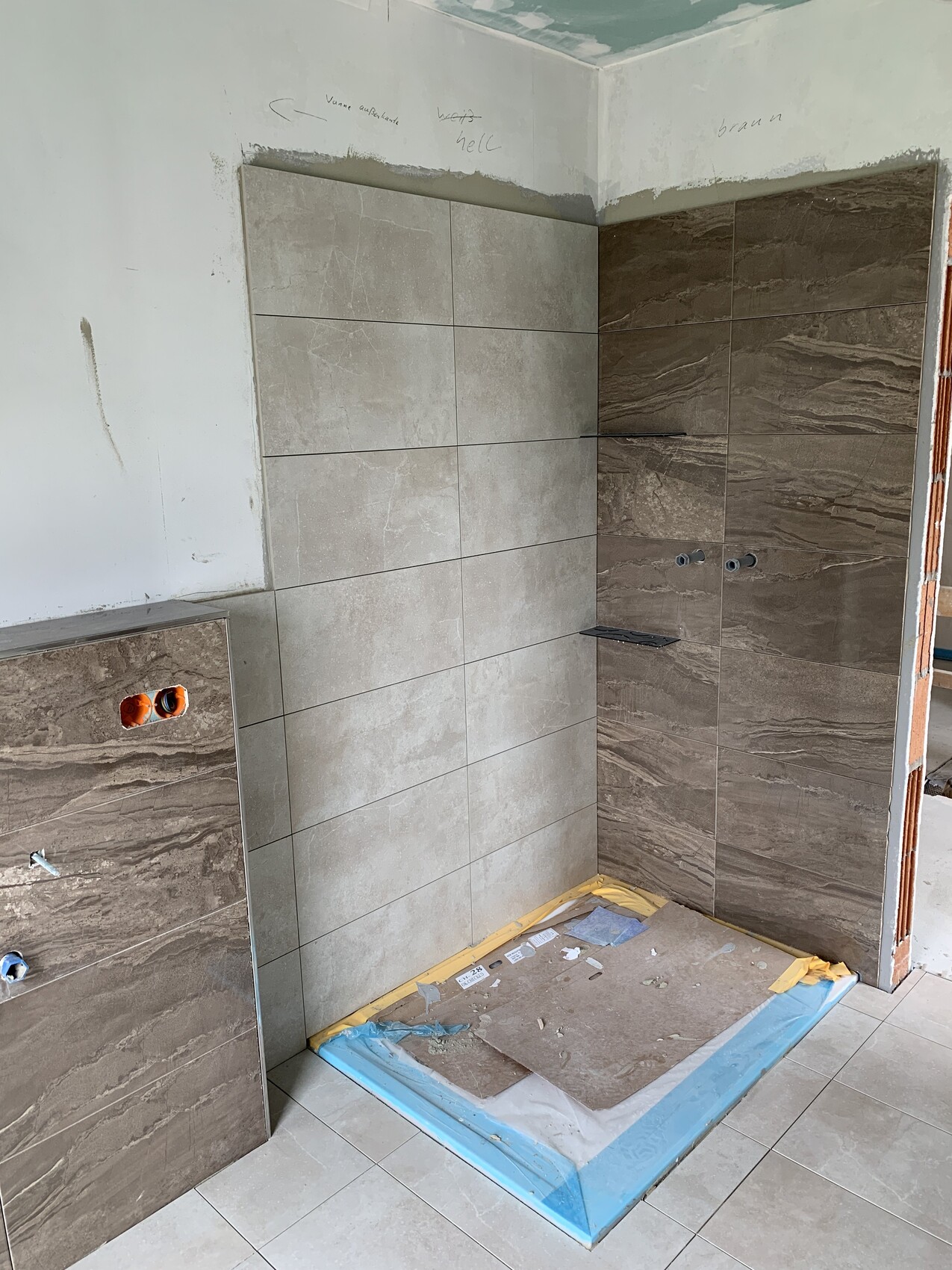 Dusche im Bau mit hellgrauen und braunen Fliesen, Boden geschützt, Rohrleitungen sichtbar.