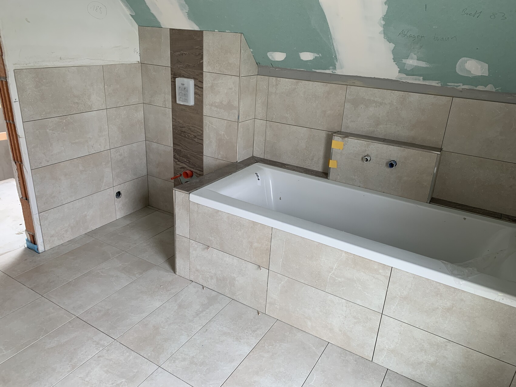 Badezimmer im Renovierungszustand: beige Fliesen, Badewanne, freiliegende Wasseranschlüsse an der Wand.