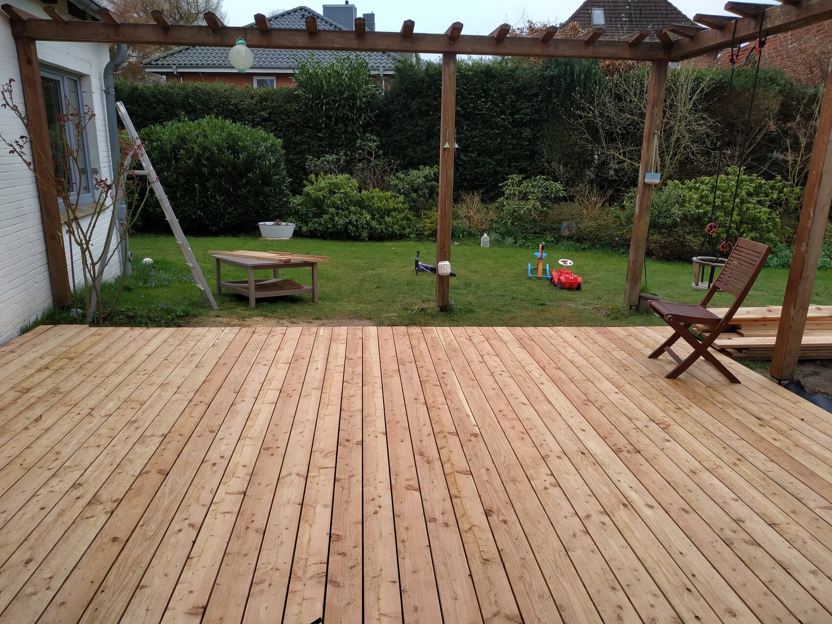 Garten mit Holzdeck und Pergola, rechts Holzstuhl, Spielzeugauto im Gras, grüne Hecke hinten.