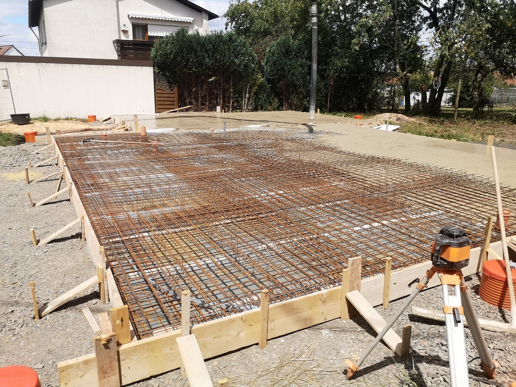 Fundamentbaustelle mit Bewehrungsstahlgitter, Holzformrahmen und Laser; Haus im Hintergrund.