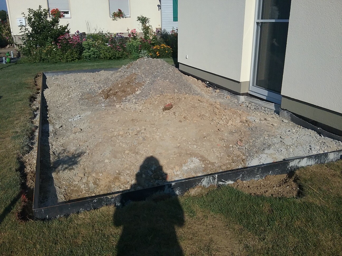 Grundstück vor Haus: Kies, Erdreich und Sandhügel; Garten mit Blumen im Hintergrund.