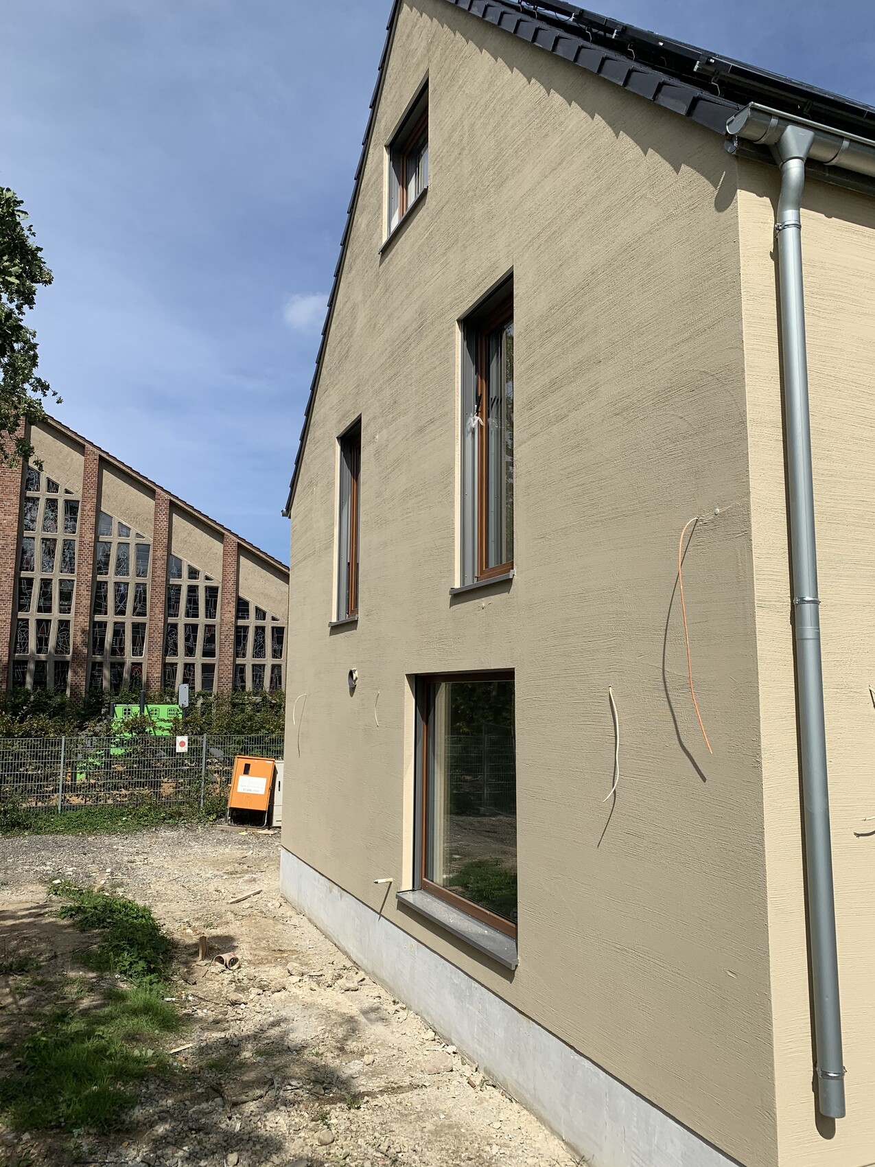 Beiger Neubau mit drei Fenstern; Baustelle davor, orange Kiste und Zaun, Hintergrund Gebäude.