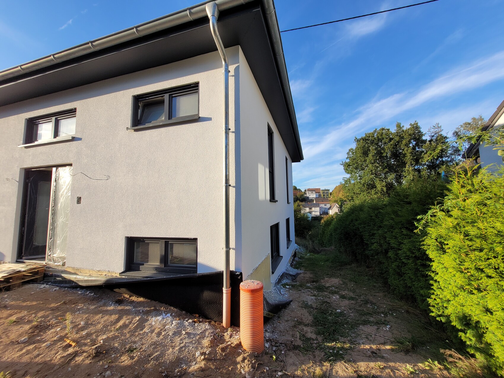 Weißes Haus im Bau mit Erdarbeiten, orange Drainagerohr am Fundament, Garten rechts.