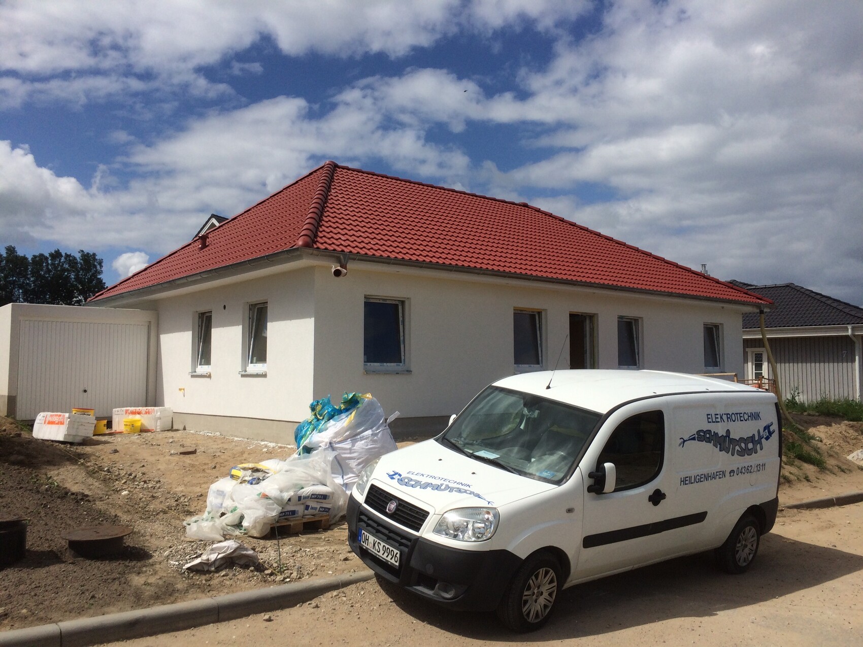 Weißes Einfamilienhaus mit rotem Ziegeldach, Baustellenmaterialien und Lieferwagen davor.