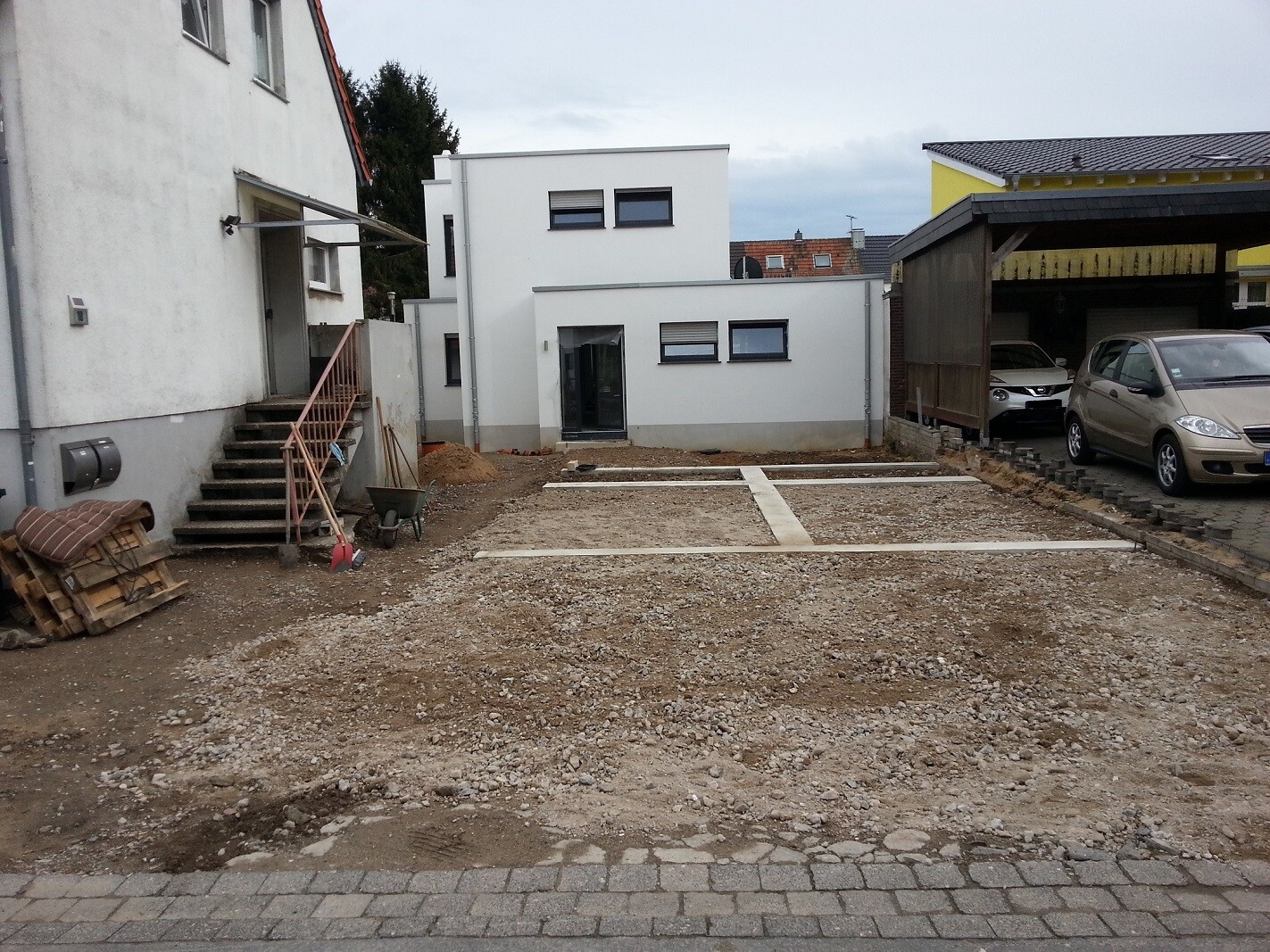 Baustelle vor modernem weißen Haus, Kiesboden, linke Treppe, rechts Carport mit Auto.