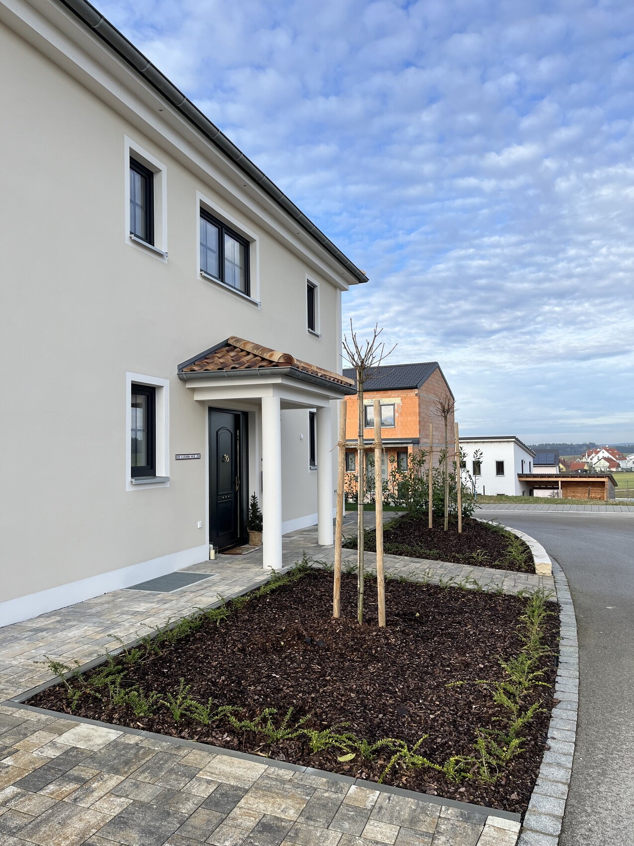 Neues beigees Mehrfamilienhaus mit Eingangspodest, weißen Säulen und gepflastertem Vorplatz.