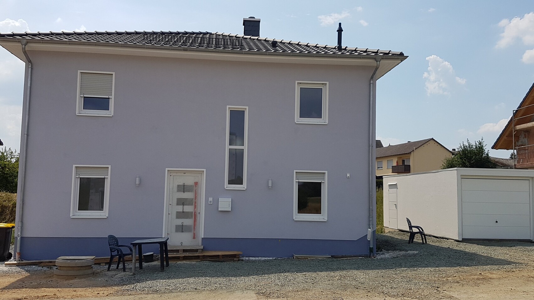 Zweistöckiges hellgraues Haus mit weißen Fenstern; Vordergrund mit Tisch und Stuhl.