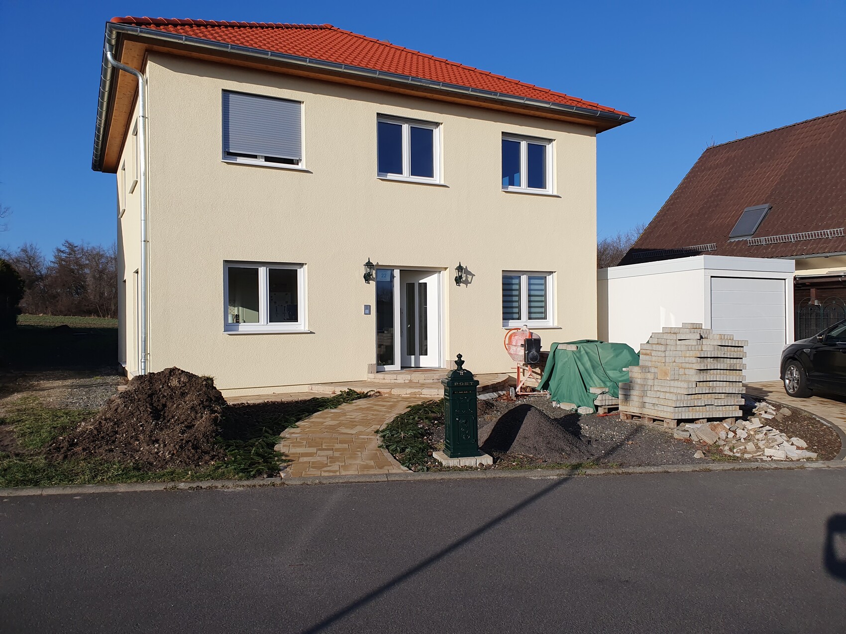 Zweistöckiges beiges Haus mit rotem Dach; Bauarbeiten vor Eingang, Materialstapel im Garten.