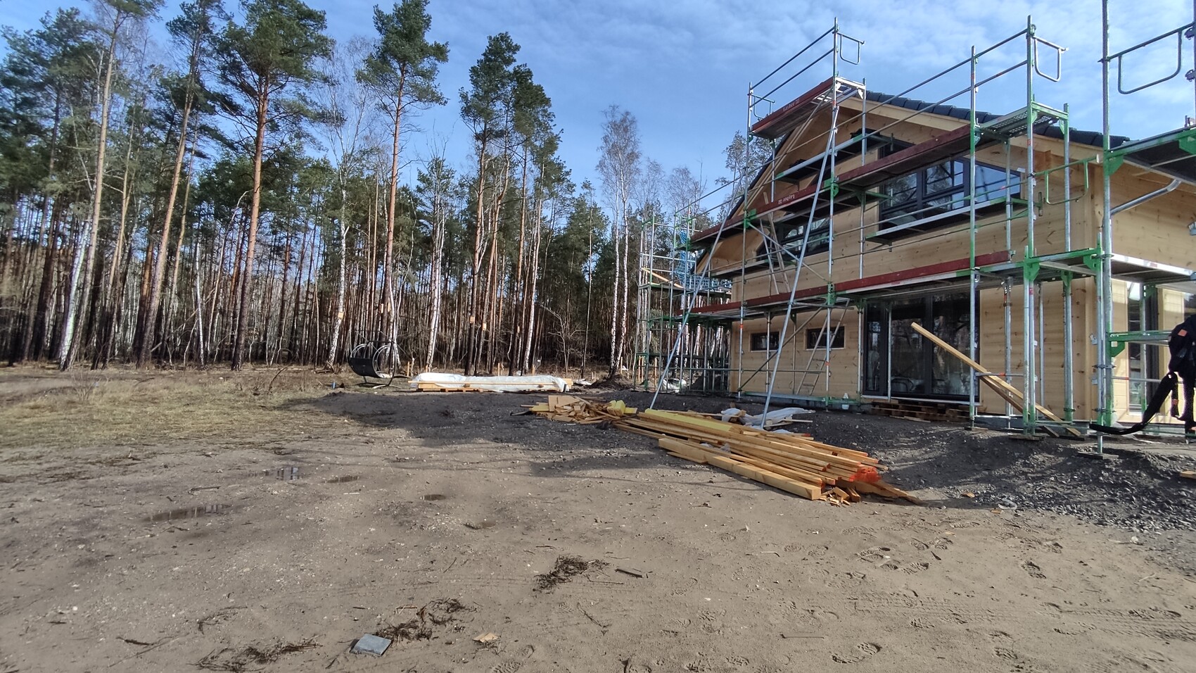 Holzhaus im Bau mit Gerüst am Waldrand; Holzbretter liegen auf dem Boden.